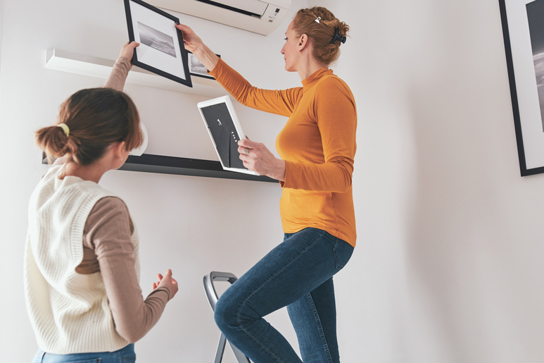 A mother stands on a step ladder to hang framed photographs on floating shelves as her daughter hands her another frame.