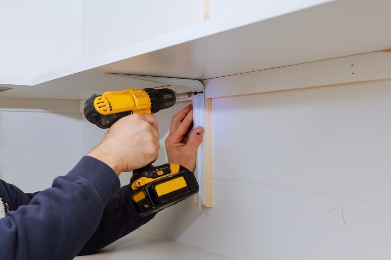 A man uses a yellow cordless drill to install a white metal bracket, securing a white particleboard shelf with a screw.