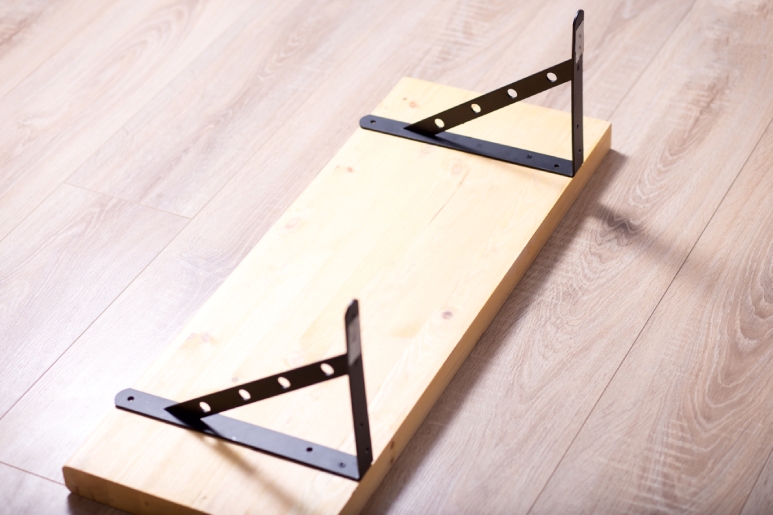 A wood shelf with two black metal brackets with triangular gussets lying on the hardwood floor.