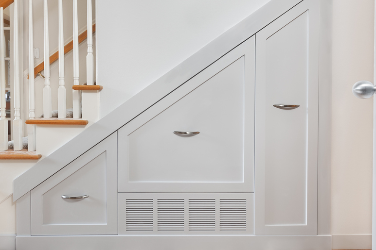 A view of a white staircase with wooden accents and the wall and space underneath it which includes pullout cabinets.