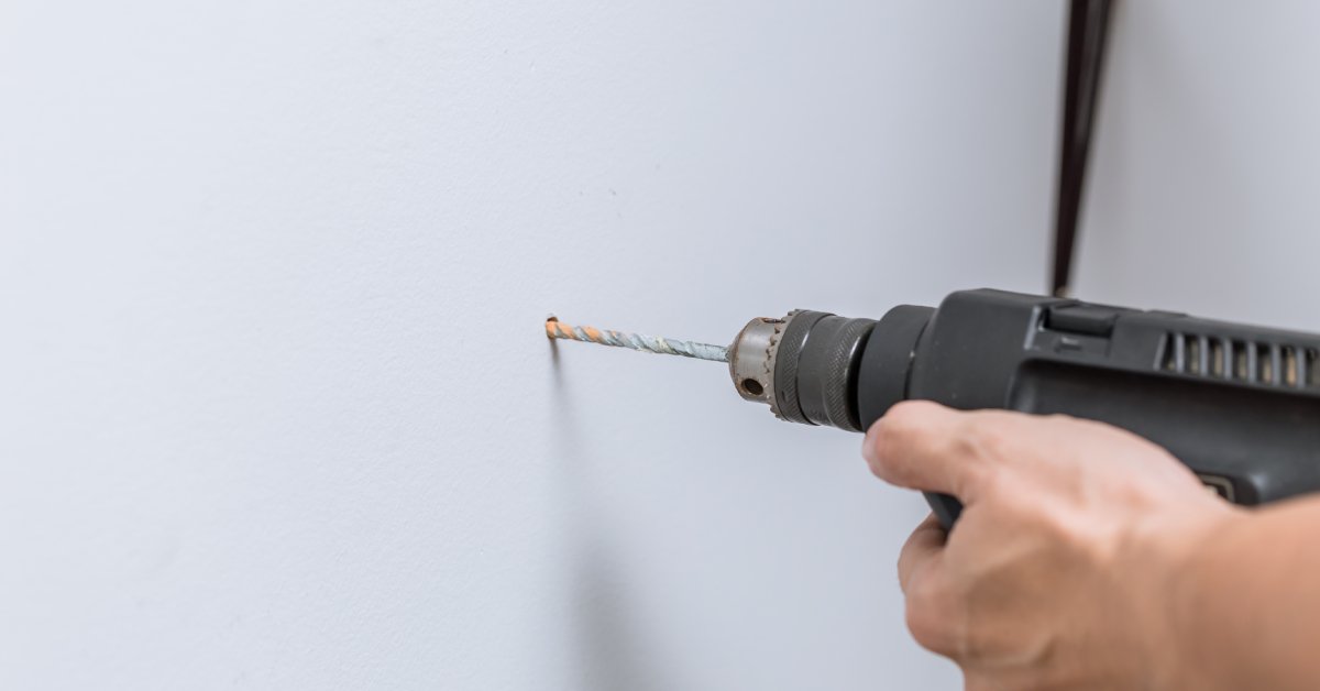 The closeup image of a person's hand holding a drill they use to insert a hole into a plain white wall.