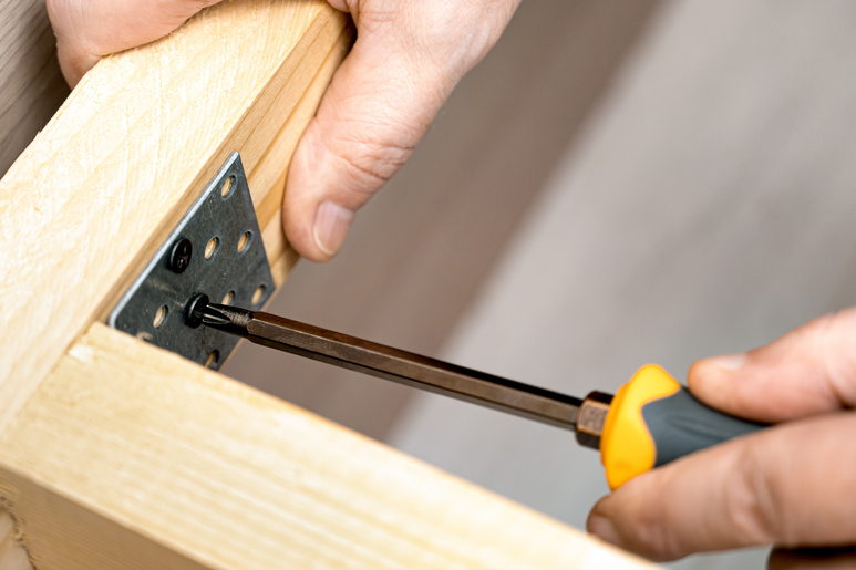 A person holds a wooden frame with their right hand and uses their left hand to tighten a screw into a metal bracket.